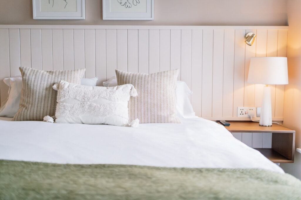 Neatly made bed with white bedding and beige textured pillows against a white paneled headboard, beside a wooden nightstand and lamp.
