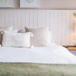 Neatly made bed with white bedding and beige textured pillows against a white paneled headboard, beside a wooden nightstand and lamp.