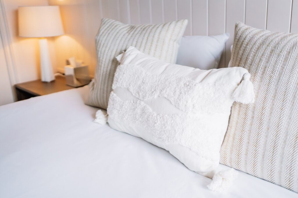 Bed made with white sheets and decorative white textured pillow, flanked by beige striped cushions in a neutral bedroom.