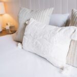 Bed made with white sheets and decorative white textured pillow, flanked by beige striped cushions in a neutral bedroom.