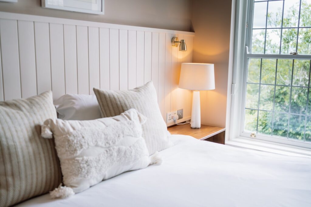 Cozy bedroom scene: white bedding with beige and textured pillows, a wooden nightstand, and a lit lamp by a large window.