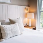 Cozy bedroom scene: white bedding with beige and textured pillows, a wooden nightstand, and a lit lamp by a large window.