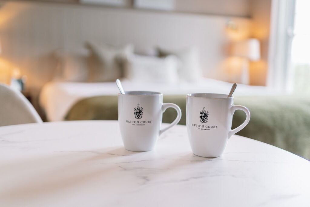 Two white mugs with a crest logo on a marble table in a cozy hotel room setting.