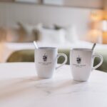 Two white mugs with a crest logo on a marble table in a cozy hotel room setting.