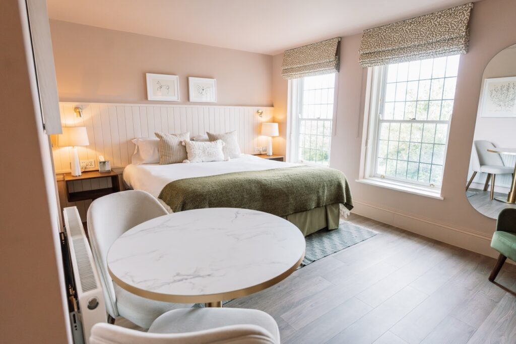 Bright hotel room with a king bed centered on a paneled wall, two bedside lamps, and a round marble-topped table by large windows.