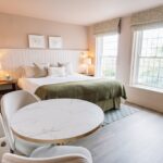 Bright hotel room with a king bed centered on a paneled wall, two bedside lamps, and a round marble-topped table by large windows.